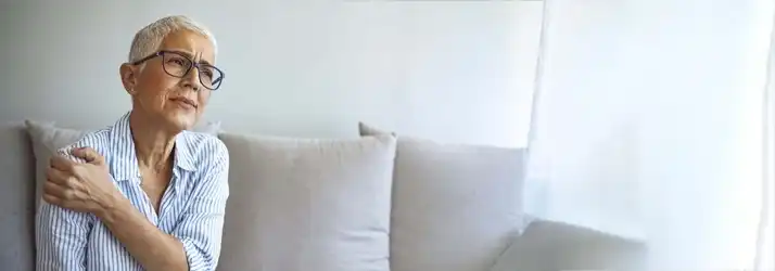 Person sitting on a couch and holding one shoulder, illustrating shoulder pain, joint discomfort, or musculoskeletal issues